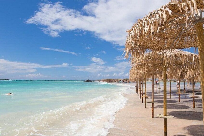 A beach with clear turquoise water, straw umbrellas, and a sandy shore. 1