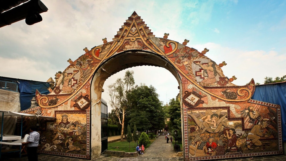 An ornate archway with intricate designs and figures, leading into a pathway surrounded by greenery. 1