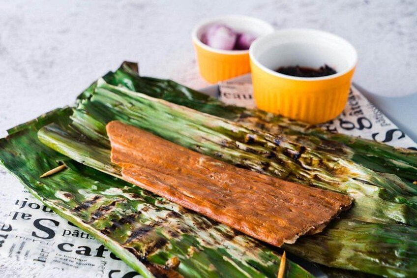 A grilled meat on banana leaves with two small bowls of sauce. 1