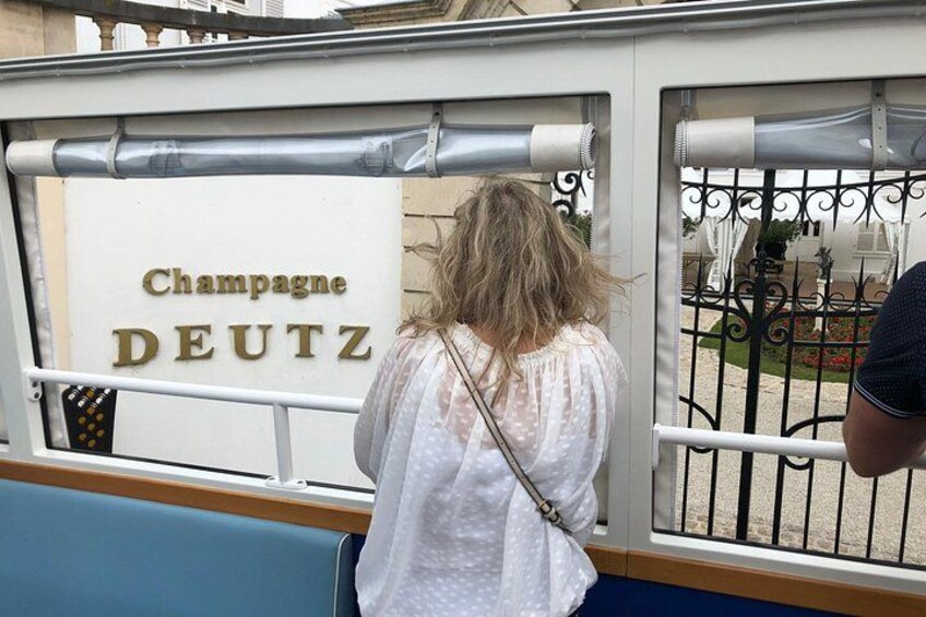 A person in a white dress standing near a gate with the sign 'Champagne DEUTZ'. 1