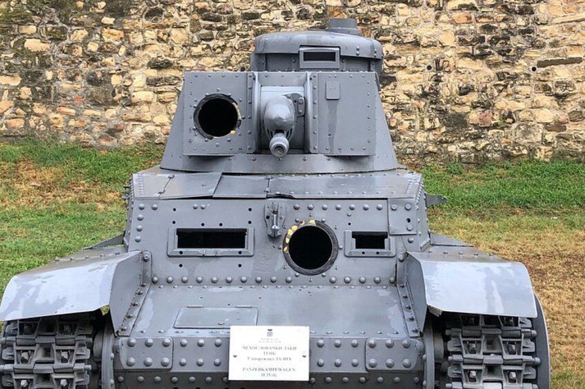 A military tank with two gun ports and a mounted machine gun, parked in front of a stone wall. 3