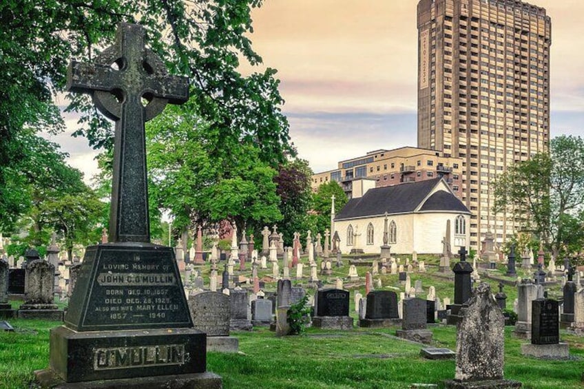 En kirkegård med et markant korsmonument, en kirke og en høj bygning i baggrunden. 2