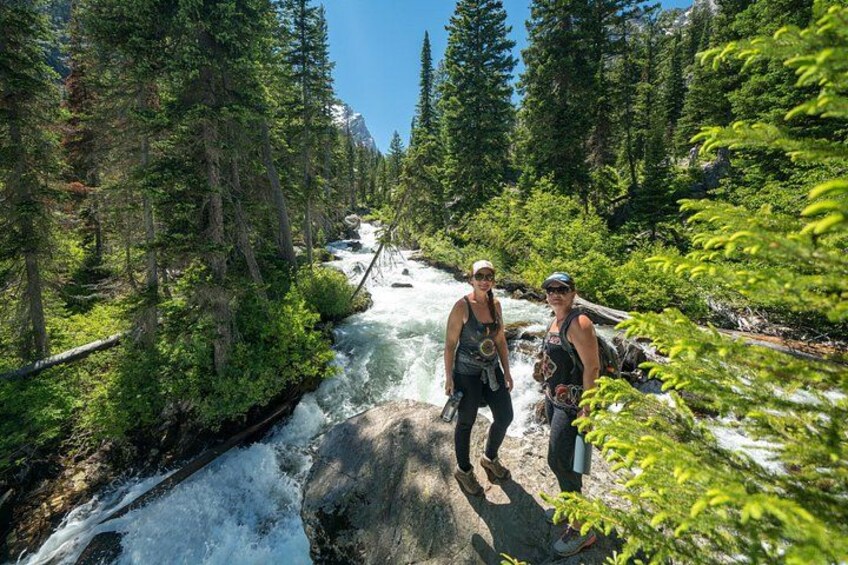 Two people hiking near a river in a forested area with snow on the ground. 3