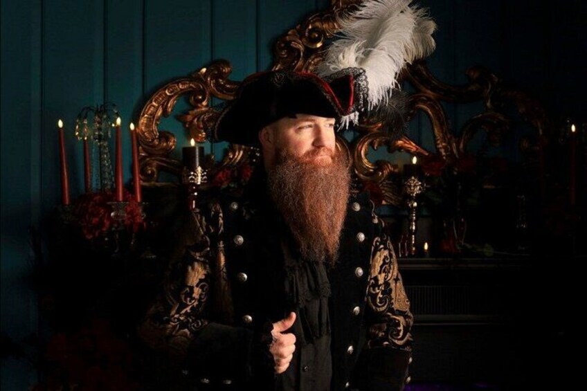 A person in ornate historical attire with a feathered hat, standing in front of a dark background with candles and a decorative chair. 4