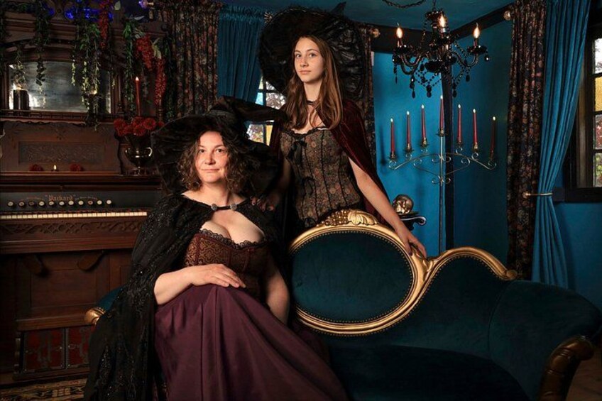 Two individuals in period costumes seated on a blue sofa in a room with a chandelier, a piano, and decorative elements. 5