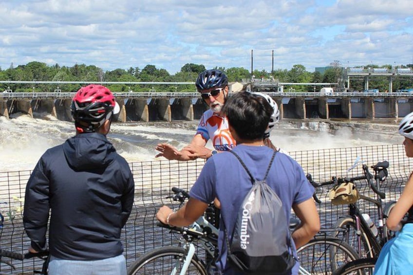 Un groupe de personnes se tient près d’une clôture, avec des vélos, et on aperçoit un pont et une étendue d’eau en arrière-plan. 4