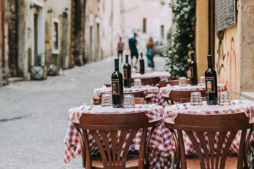 Des tables installées à l’extérieur pour les repas, recouvertes de nappes à carreaux et ornées de bouteilles de vin, dans une ruelle bordée de bâtiments des deux côtés. 1