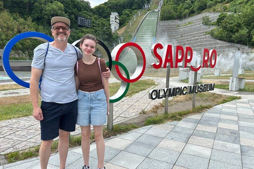 A man and a woman standing in front of the Sapporo Olympic Museum sign with Olympic rings and a stadium in the background. 2