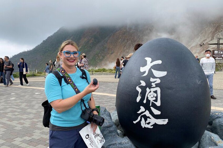 A person with a camera and a large black stone with white writing, standing in a mountainous area with other people in the background. 4