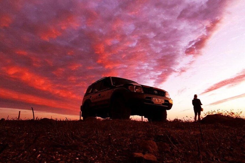 Un véhicule est garé dans un champ, et une personne se tient à côté, sous un ciel flamboyant au coucher du soleil. 3
