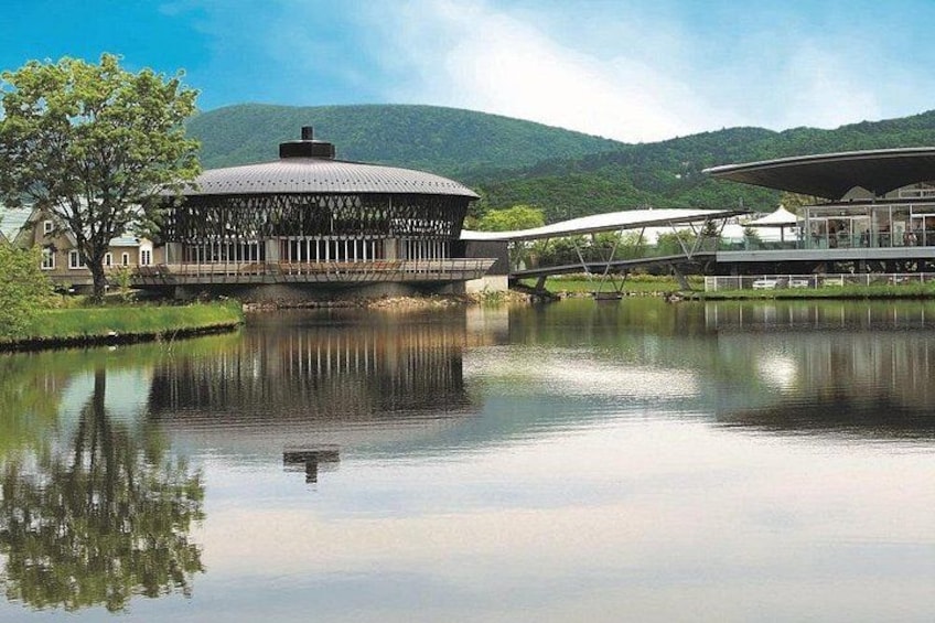 A modern building with a circular roof near a calm lake, surrounded by greenery and hills. 1