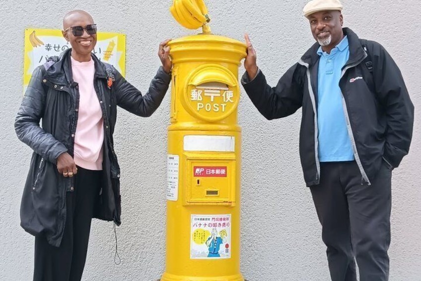 Two people standing next to a yellow post box with Japanese text. 5