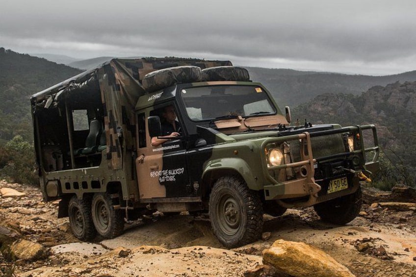 A military vehicle with a camouflage pattern, equipped with a roof rack, parked on a rocky terrain with a mountainous backdrop. 1