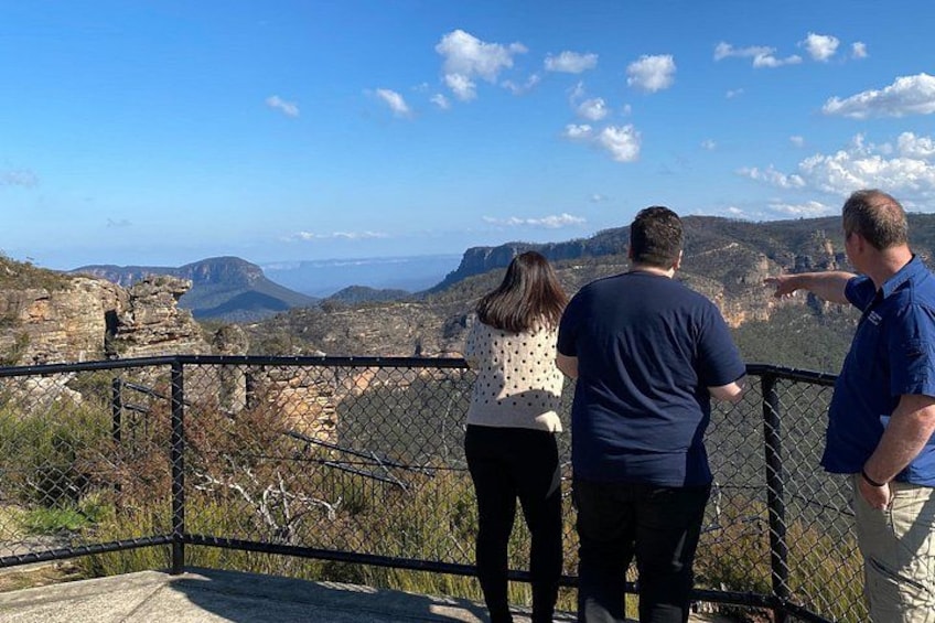 Three people standing on a viewing platform overlooking a mountainous landscape with ruins and a clear sky. 4