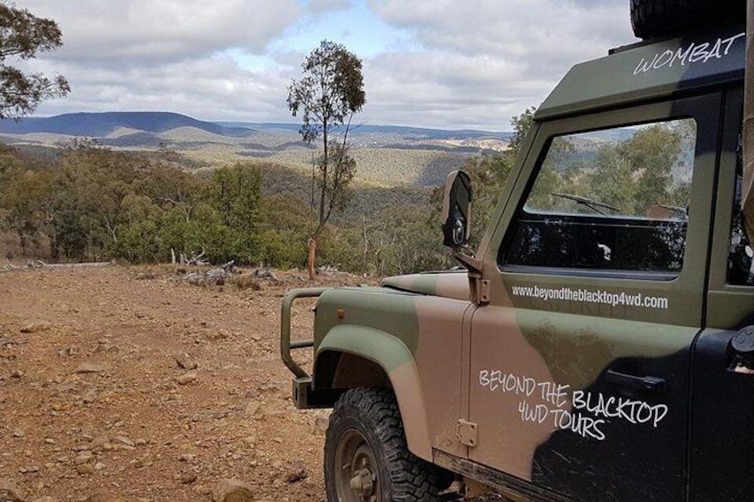 A beige off-road vehicle with 'Beyond the Blacktop Tours' written on its side, parked on a dirt road with a scenic view of hills and sparse vegetation in the background. 2
