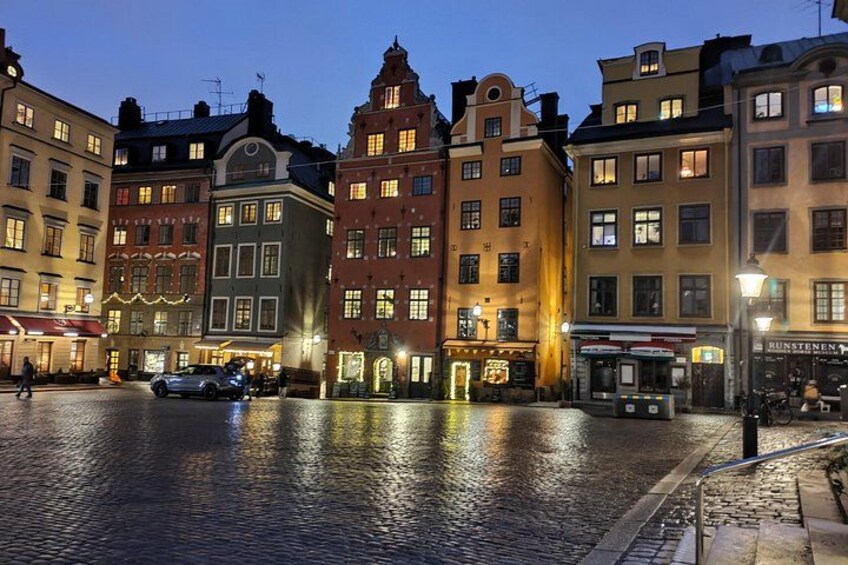En kullerstensgata i skymningen med historiska byggnader, en parkerad bil och en busshållplats. 1