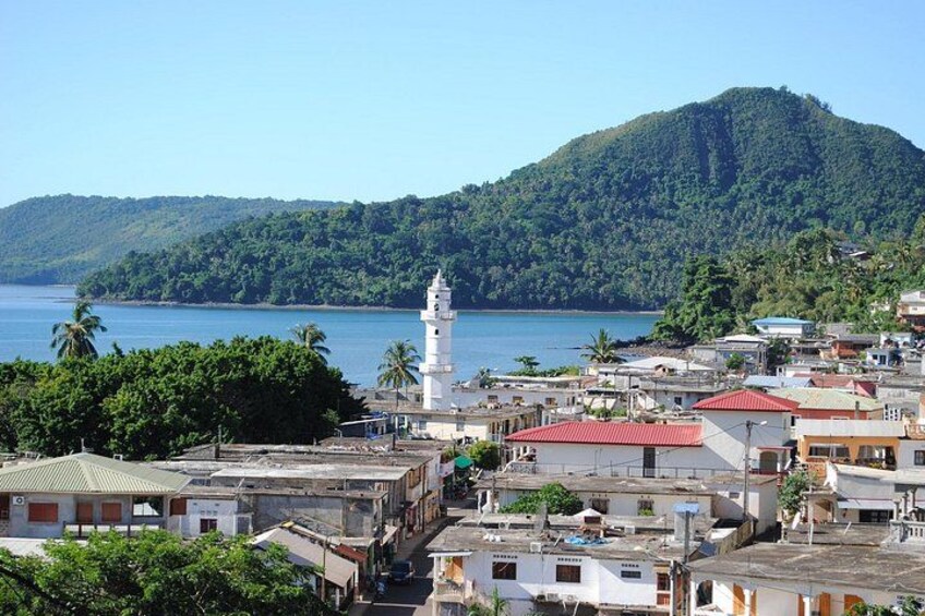 Un pueblo costero con un faro blanco destacado, vegetación densa y una montaña al fondo. 1