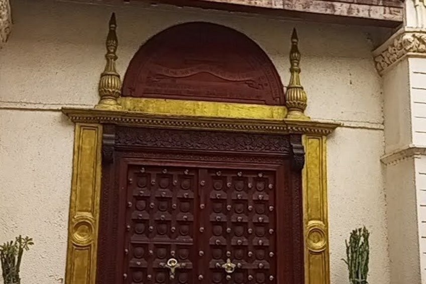 Una puerta de madera grande con tallas intrincadas y un cerrojo de bronce, flanqueada por columnas decorativas y una pequeña planta. 4