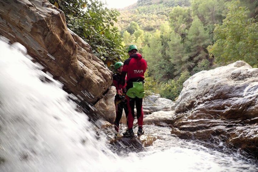 Two people in red jackets standing on a rocky ledge by a waterfall. 2