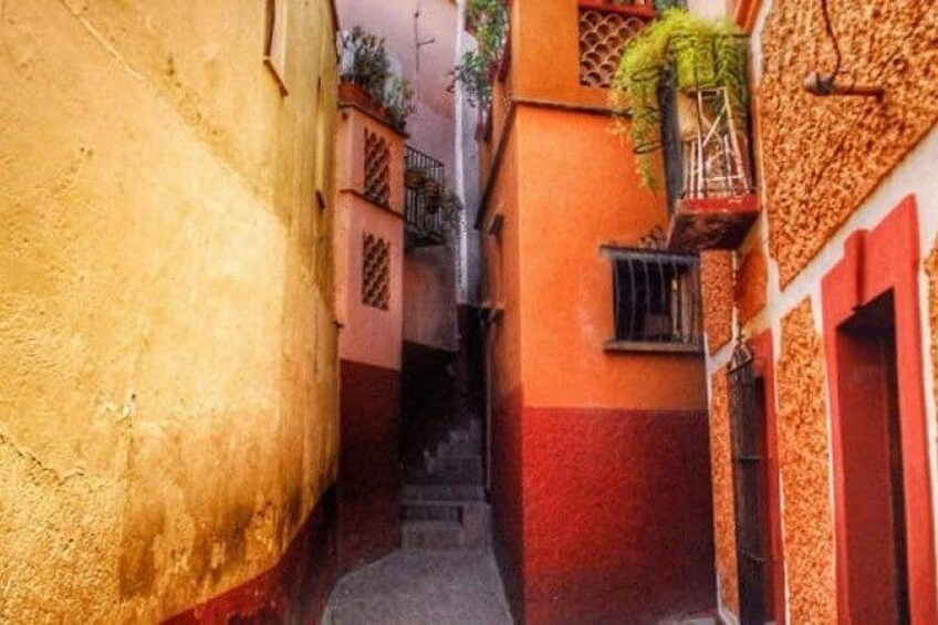 A narrow alleyway with buildings painted in warm tones, a staircase, and a small window with a plant. 2