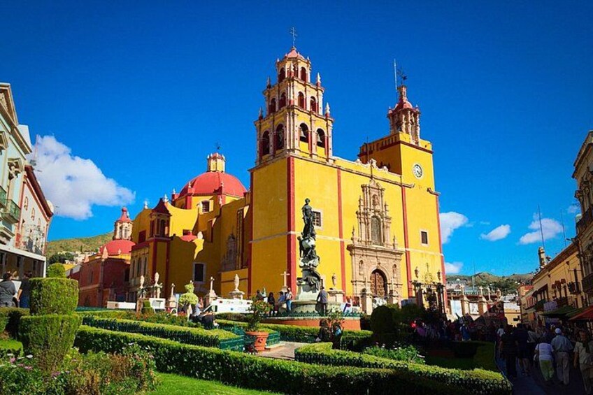 A historic yellow church with two bell towers, surrounded by well-manicured gardens and a clear blue sky. 1