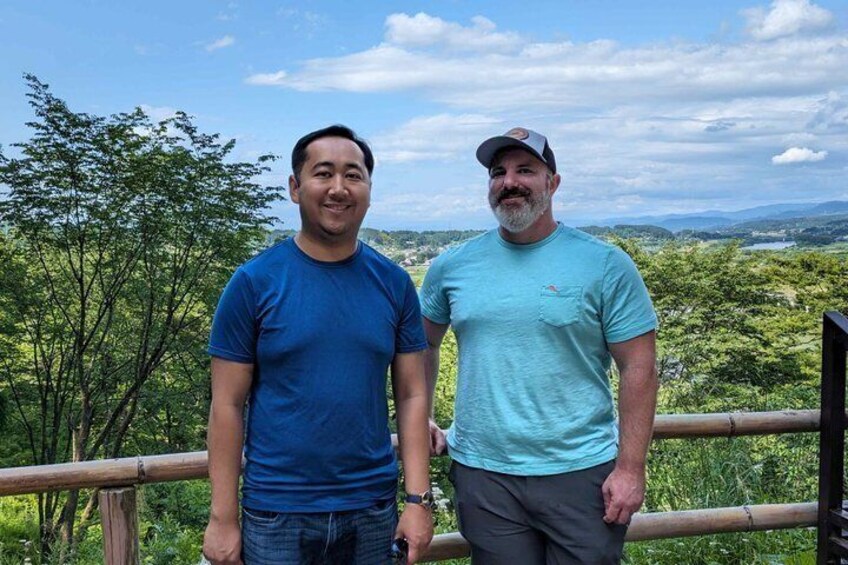 Two individuals standing on a wooden deck with a scenic view of a forested landscape and distant hills. 3