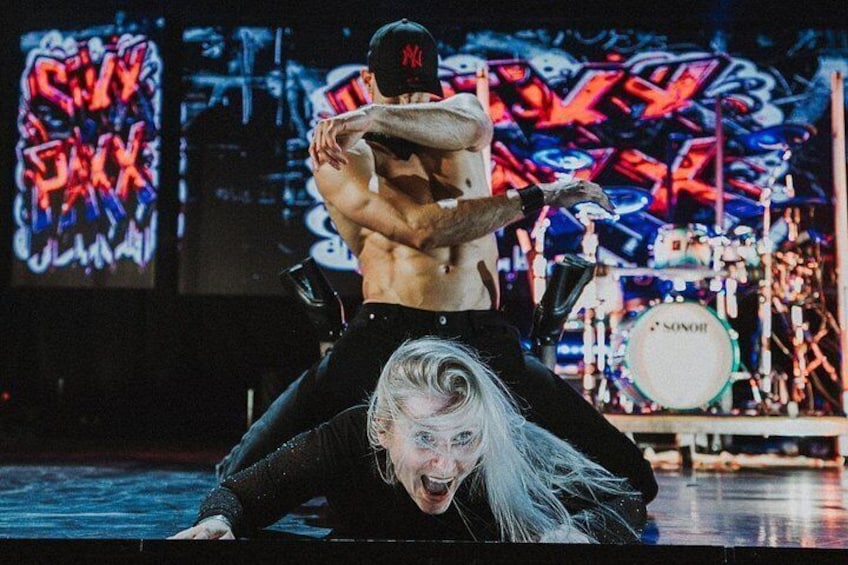 A person in a black outfit with a white wig, another person in a black cap and tank top, and a drum set in the background. 4