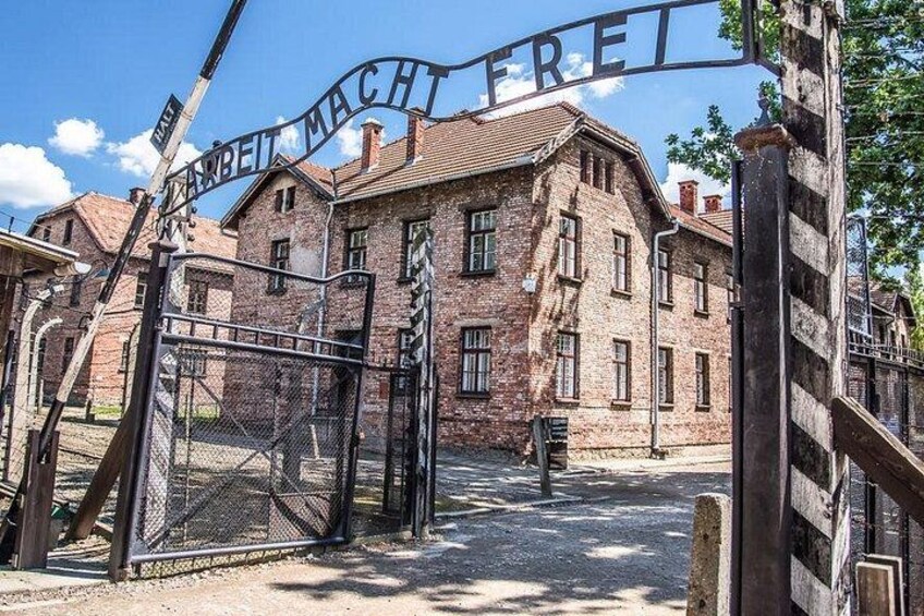 A historic building with a sign reading 'ARBEIT MACHT FREI' in front of a metal gate. 1