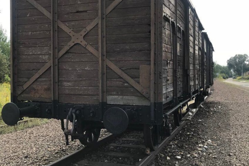 A wooden train car with a cross-brace structure, black wheels, and a gravel railway track. 2