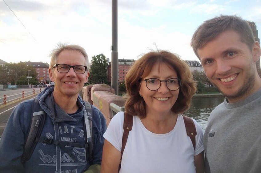 Tre personer står på en bro med en flod og bygninger i baggrunden. 2