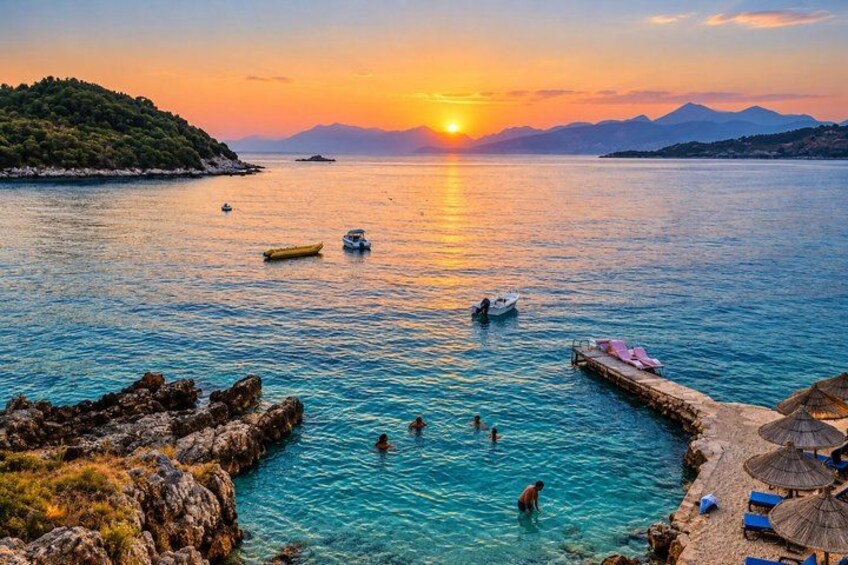 A sunset over a calm sea with boats, rocky shore, and people swimming. 2
