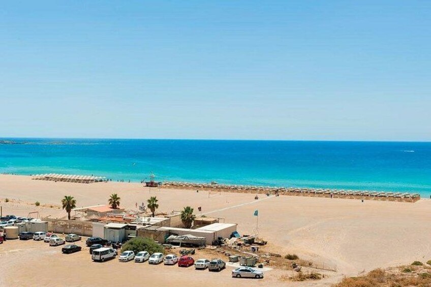 Una playa con agua azul cristalina, un estacionamiento y algunas palmeras. 1