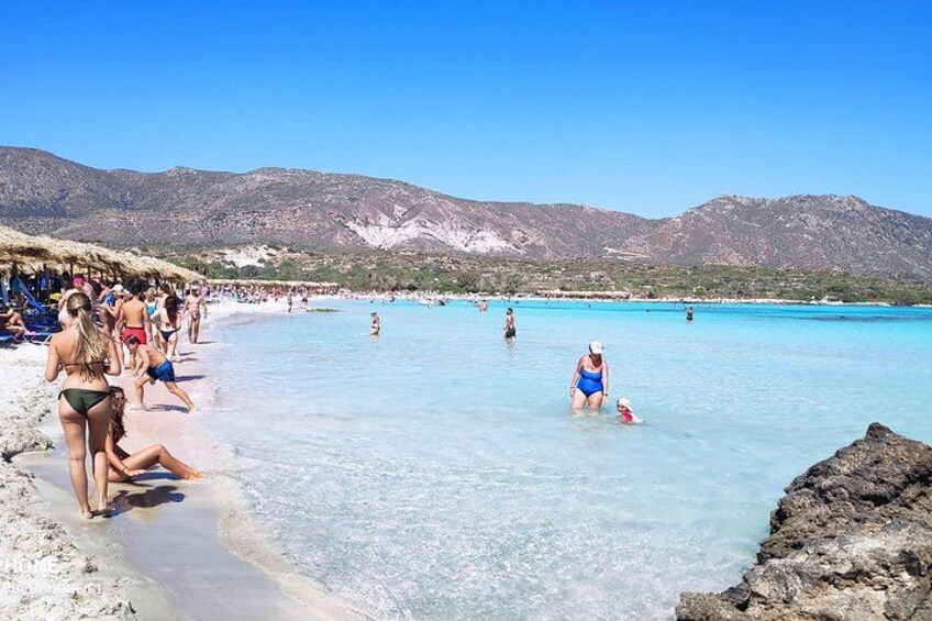 A beach with people swimming and walking, clear turquoise water, and mountains in the background. 4