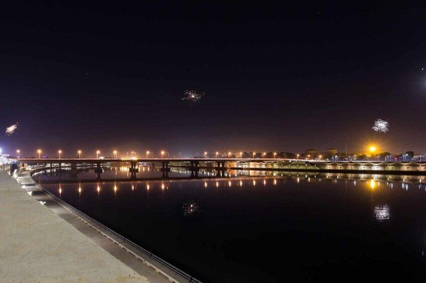 Une vue nocturne d’un front de mer avec un pont, des lumières se reflétant sur l’eau et des feux d’artifice dans le ciel. 1