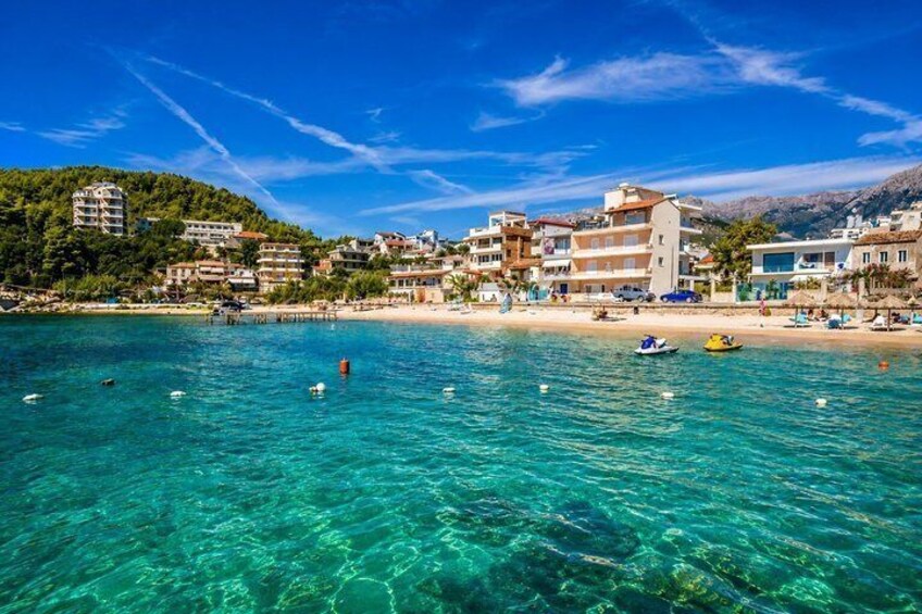 A coastal town with clear turquoise waters, a sandy beach, and buildings along the shore. 1