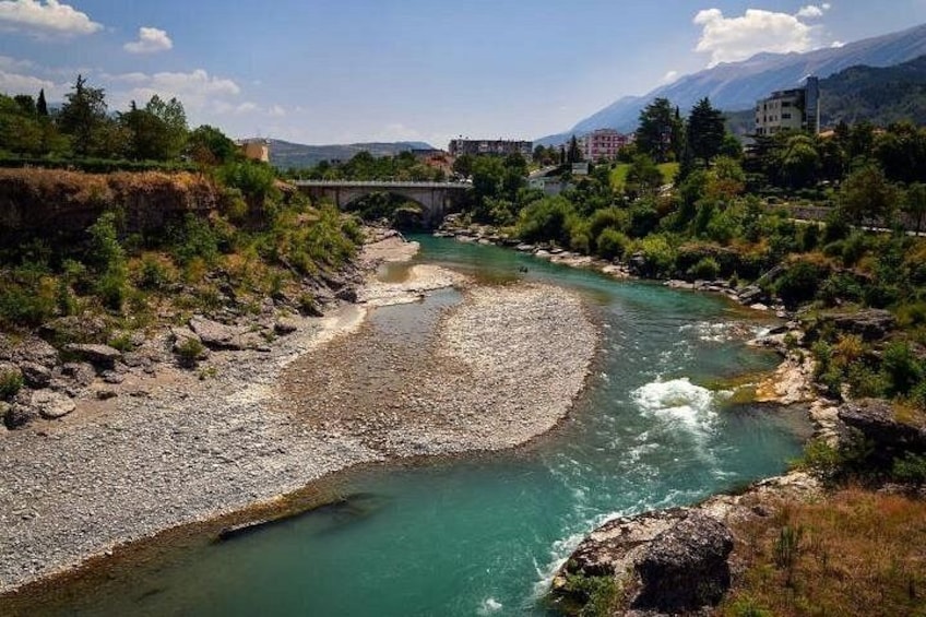 A river with a rocky riverbed, a bridge, and buildings in the background. 1