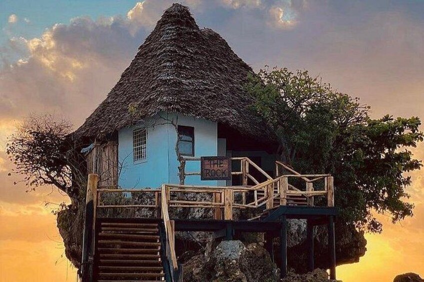 A house with a thatched roof on stilts, surrounded by trees, with a sunset in the background. 1