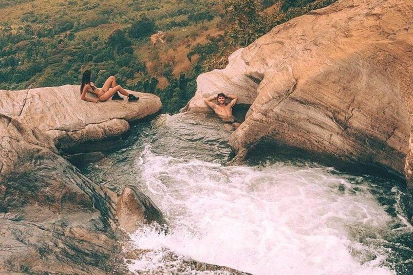 A person sitting on a rock by a flowing river, another person in the water, and a rocky landscape with greenery in the background. 5