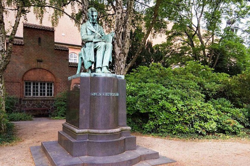 Una estatua de un hombre sentado con una placa que dice 'Soren Kierkegaard' en un jardín con un edificio de ladrillo al fondo. 4