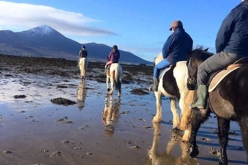 Three people riding horses on a wet beach with a mountain in the background. 1