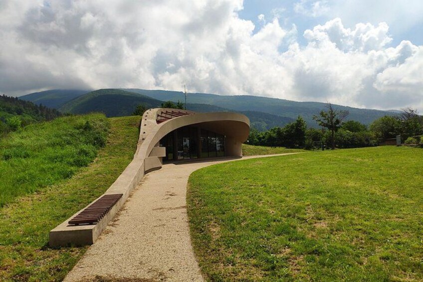 A modern building with a curved roof, surrounded by a grassy field and mountains in the background. 3