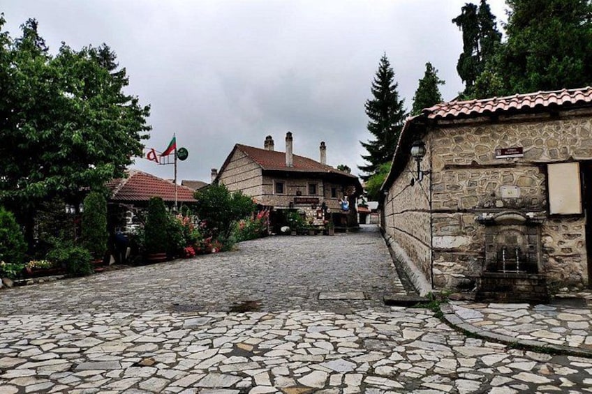 A cobblestone street leading to a stone building with a tiled roof, surrounded by trees and greenery. 1