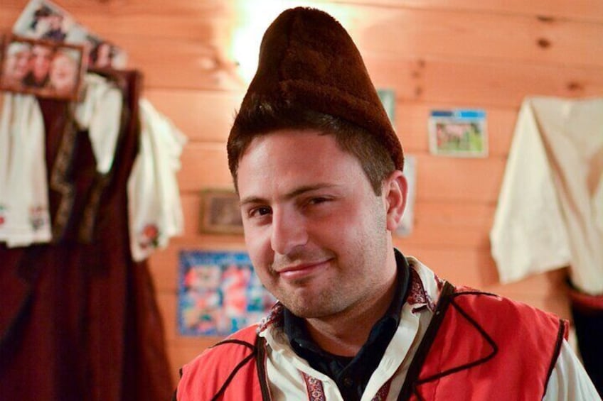 A person wearing a traditional hat and a red vest in a wooden room with clothing hanging on the wall. 3