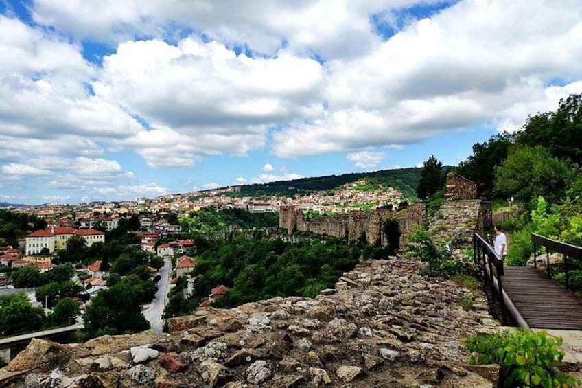 A historic stone bridge, a wooden walkway, and a cityscape with buildings and greenery under a partly cloudy sky. 2