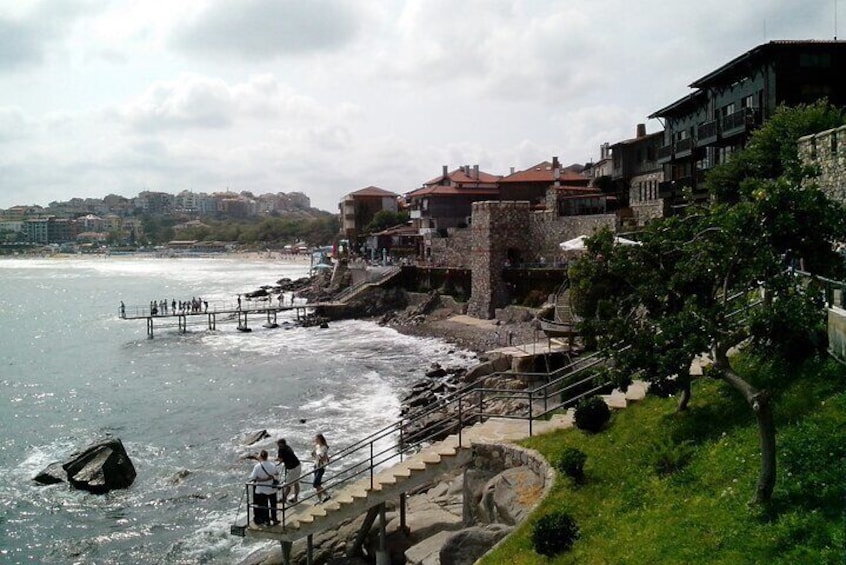 A coastal town with a stone staircase leading down to the beach, a pier with people, and buildings along the shore. 1