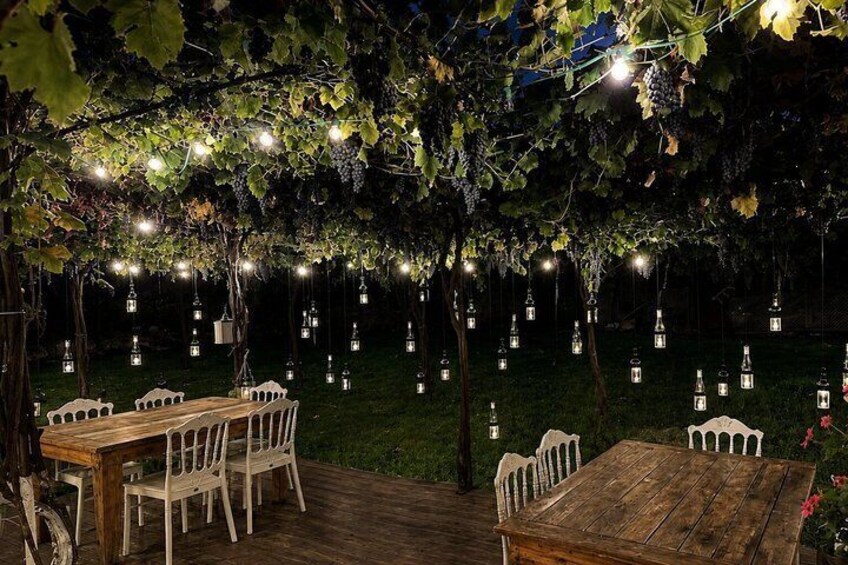Een buiten eethoek met houten tafels en stoelen, onder een pergola begroeid met wijnranken en verlicht met hangende lantaarns. 4