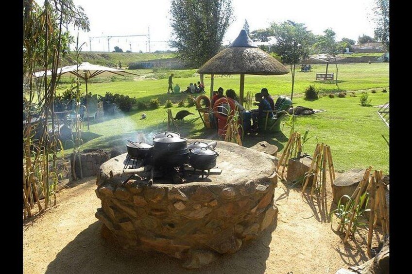 An outdoor cooking area with a stone structure, surrounded by a grassy field and a thatched-roof pavilion. 4