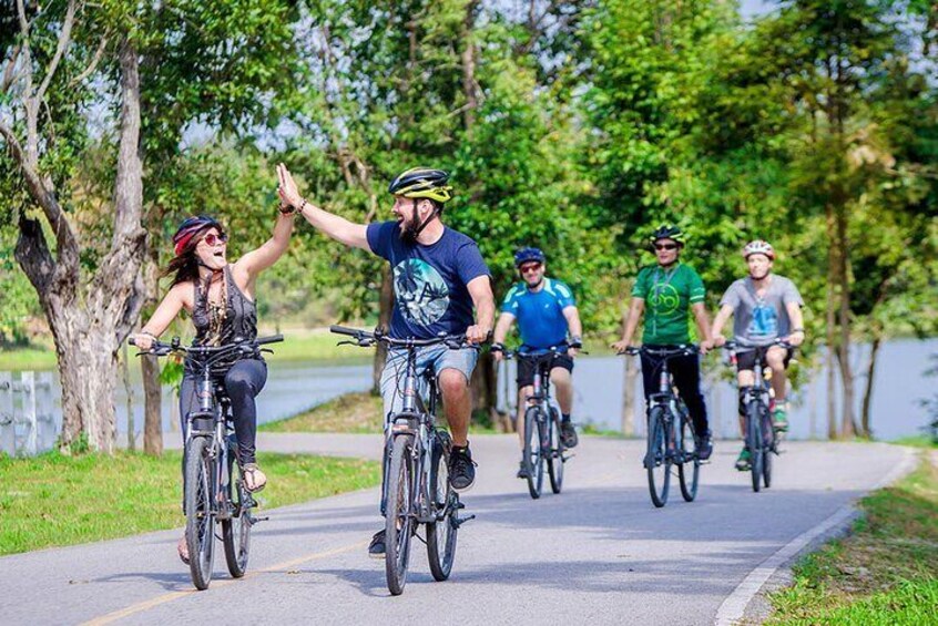 Un grupo de personas pedaleando en bicicleta por un sendero pavimentado, rodeado de árboles y un cuerpo de agua. 1