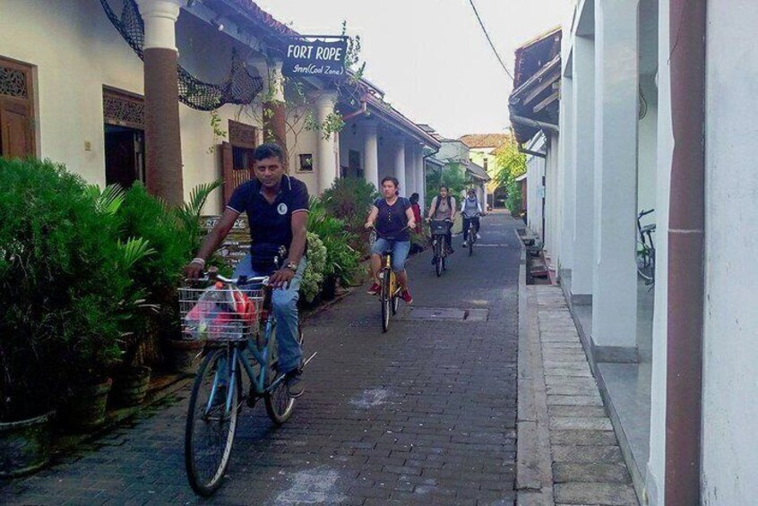 Una calle estrecha con ciclistas circulando por la izquierda, un edificio con un letrero que dice "FORT ROPE Inn (Col. Zone)" a la derecha, y follaje a lo largo de la izquierda. 5