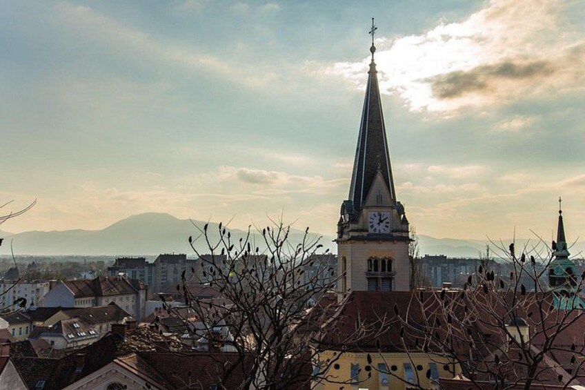 A cityscape with a prominent church tower, a mountain in the background, and leafless trees in the foreground. 3